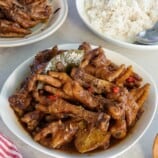 adobong paa ng manok in a white bowl with a plate of steamed rice in the background.