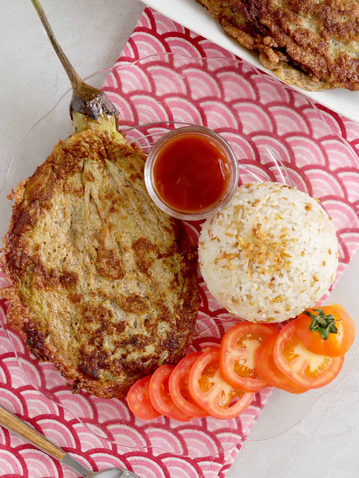 Tortang Talong on a clear glass plate with steamed rice and sliced tomatoes on the side.